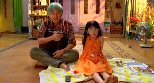 A screenshot of a young man with blue hair holding a briefcase sitting on the floor with a little girl. From the film Korean 'Sympathy for Mr. Vengeance'.