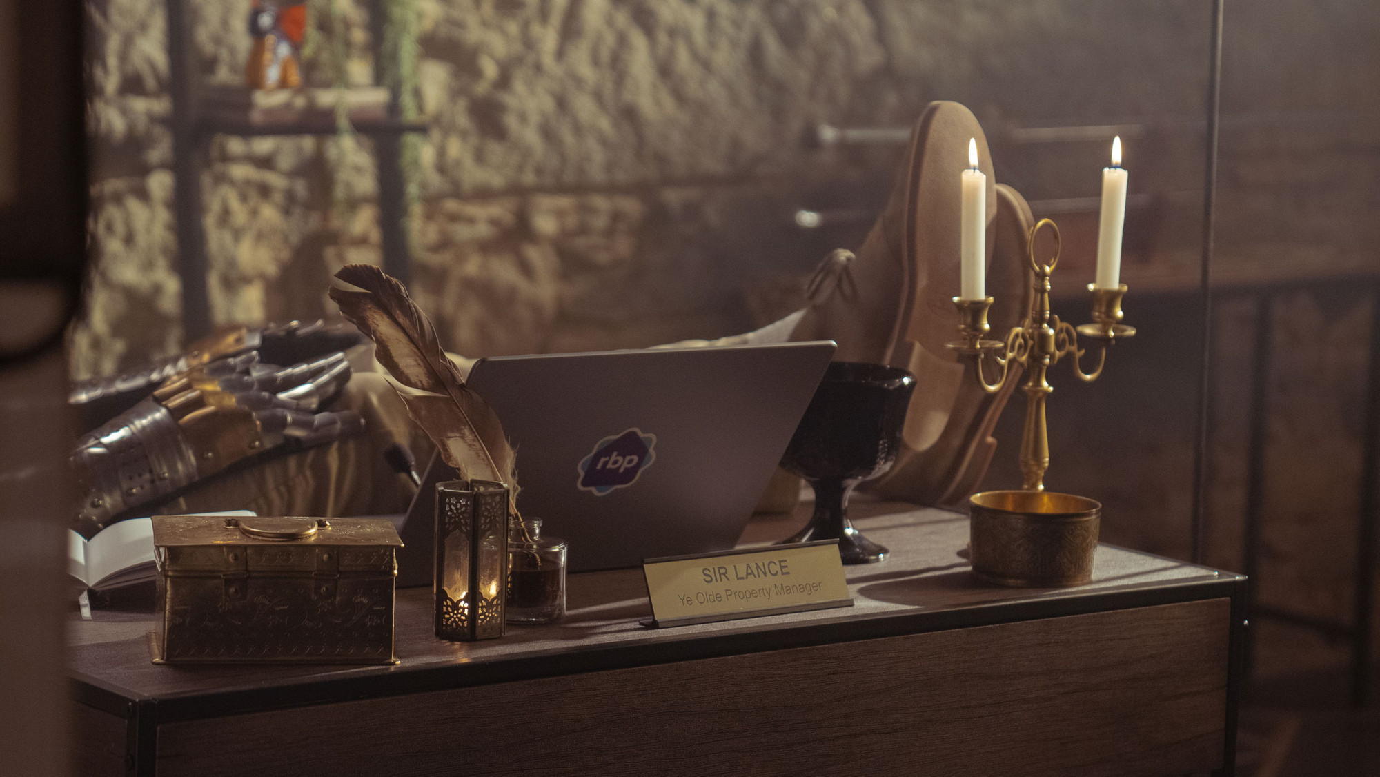 A knight’s feet propped up on a desk with a laptop and a nameplate that reads ‘Sir Lance Ye Olde Property Manager’