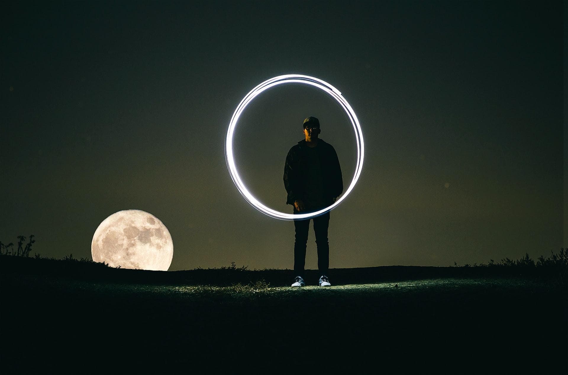 man standing on hill during full moon, painting a circle with light and long camera exposure