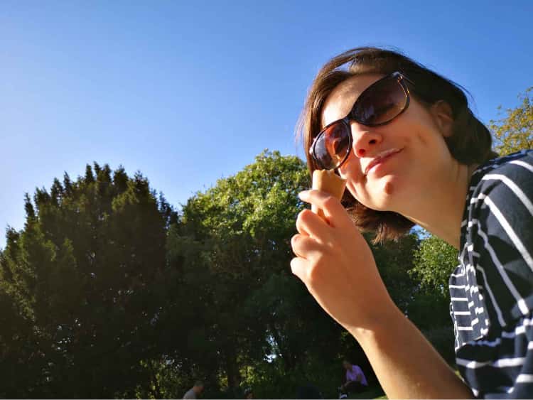 Naomi with a big smile on her face, holding an ice cream, with a bright blue sky and some green Museum Garden trees as the backdrop.