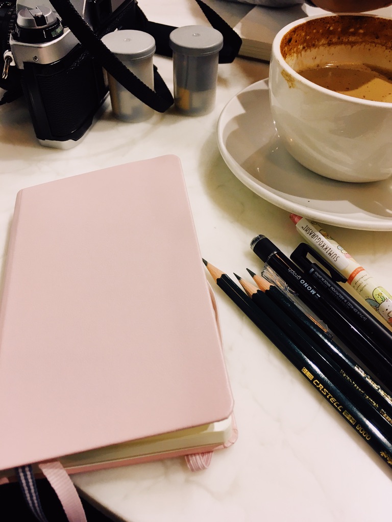 A table with a small sketchbook, an assortment of pencils, a cup of coffee, and a film camera.