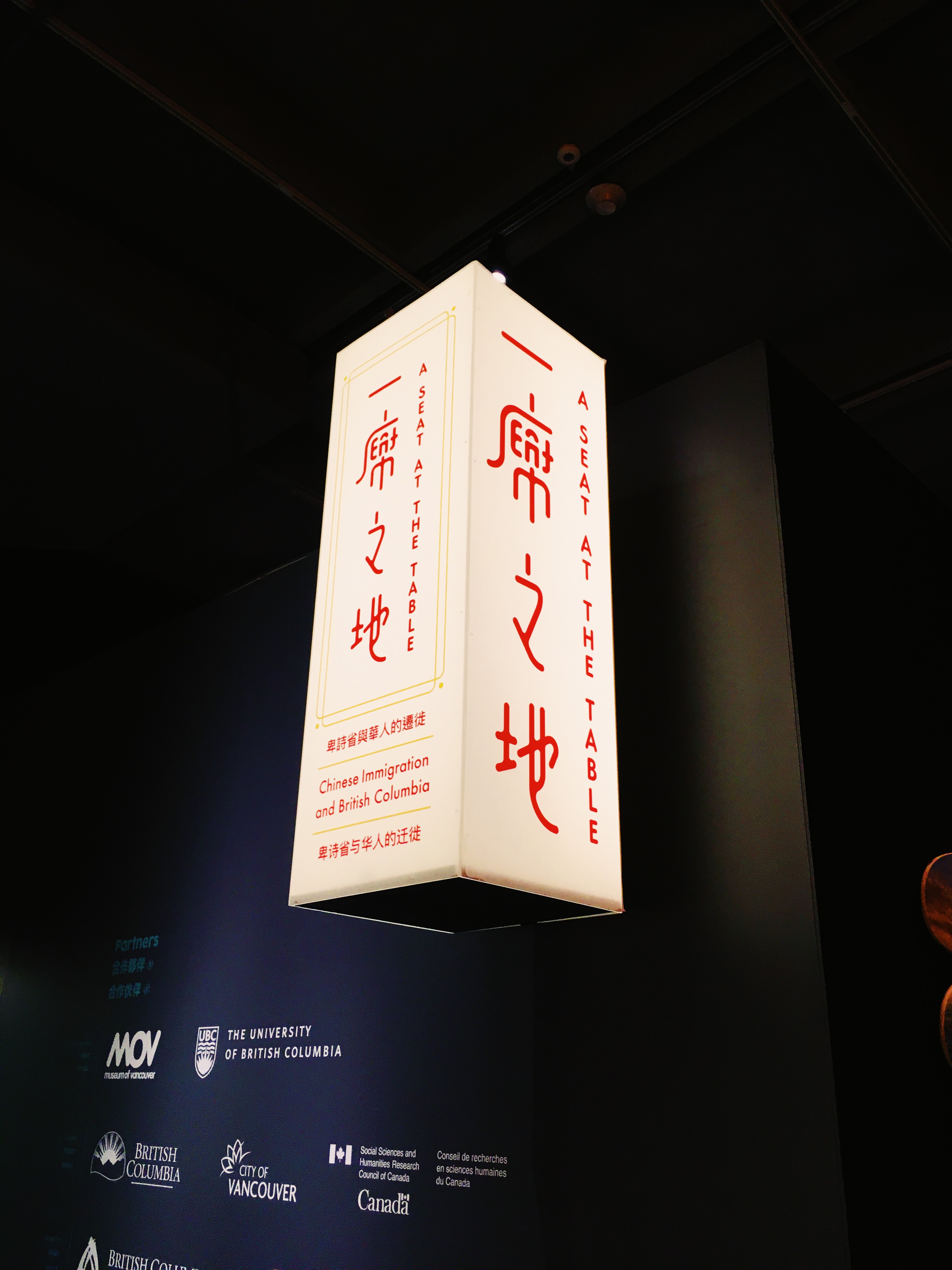 Exhibit sign that reads 'A Seat at the Table'. The sign is a tall white box with red text, in both English and Chinese.