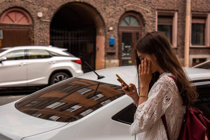 A caucasian female standing next to a silver car and looking down at her mobile device while she puts her left hand on her face.