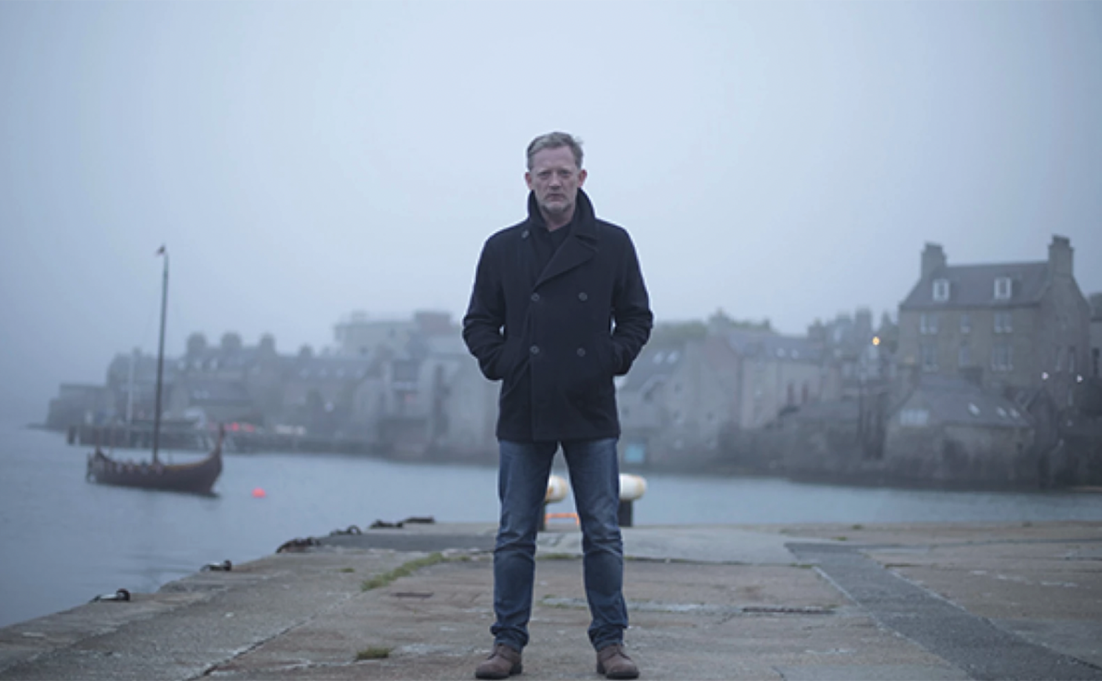  photo of actor douglas henshall in a blue coat standing on a dock by the ocean