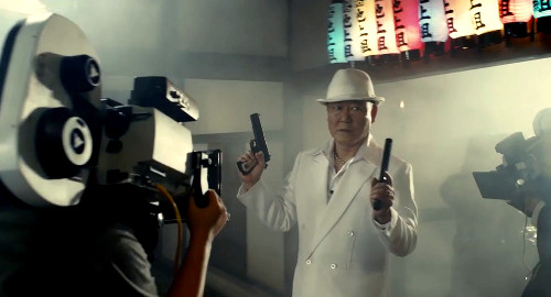 A screenshot of the film 'Why Don’t You Play in Hell?' showing cameras on a yakuza leader dressed in a white suit and hat walking into a smokey room with a pistol in each hand.