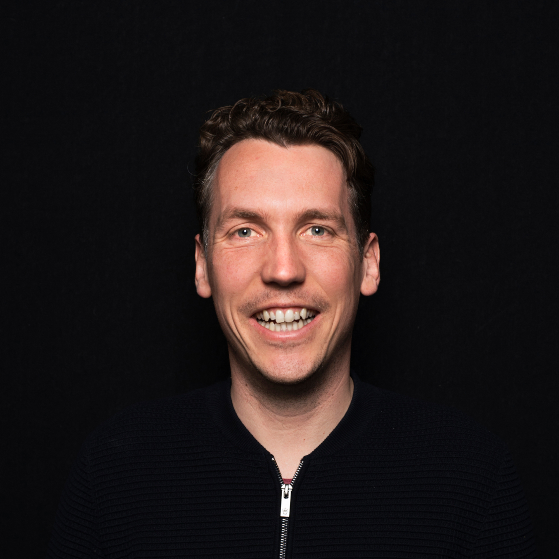 A light brown haired caucasian man smiling in the midst of a black background, wearing a black bomber jacket.