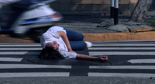A screenshot from the movie 'The Terrorizors' showing a young woman lying in a crosswalk as motorbikes drive by.