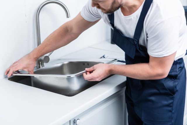 bearded repairman in working overall fixing sink in kitchen