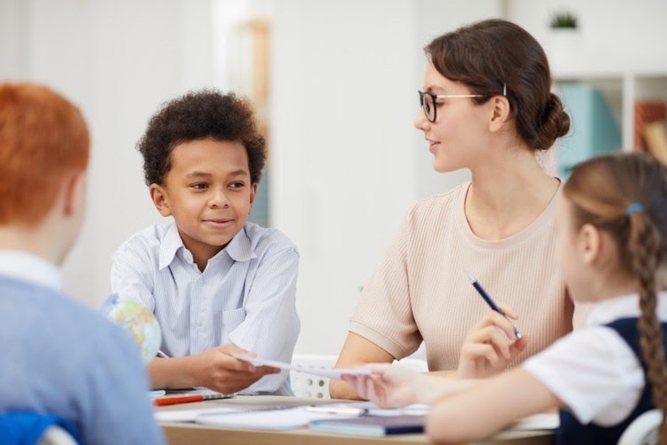A smiling elementary school teacher sits at a classroom table and leads a discussion with three students.