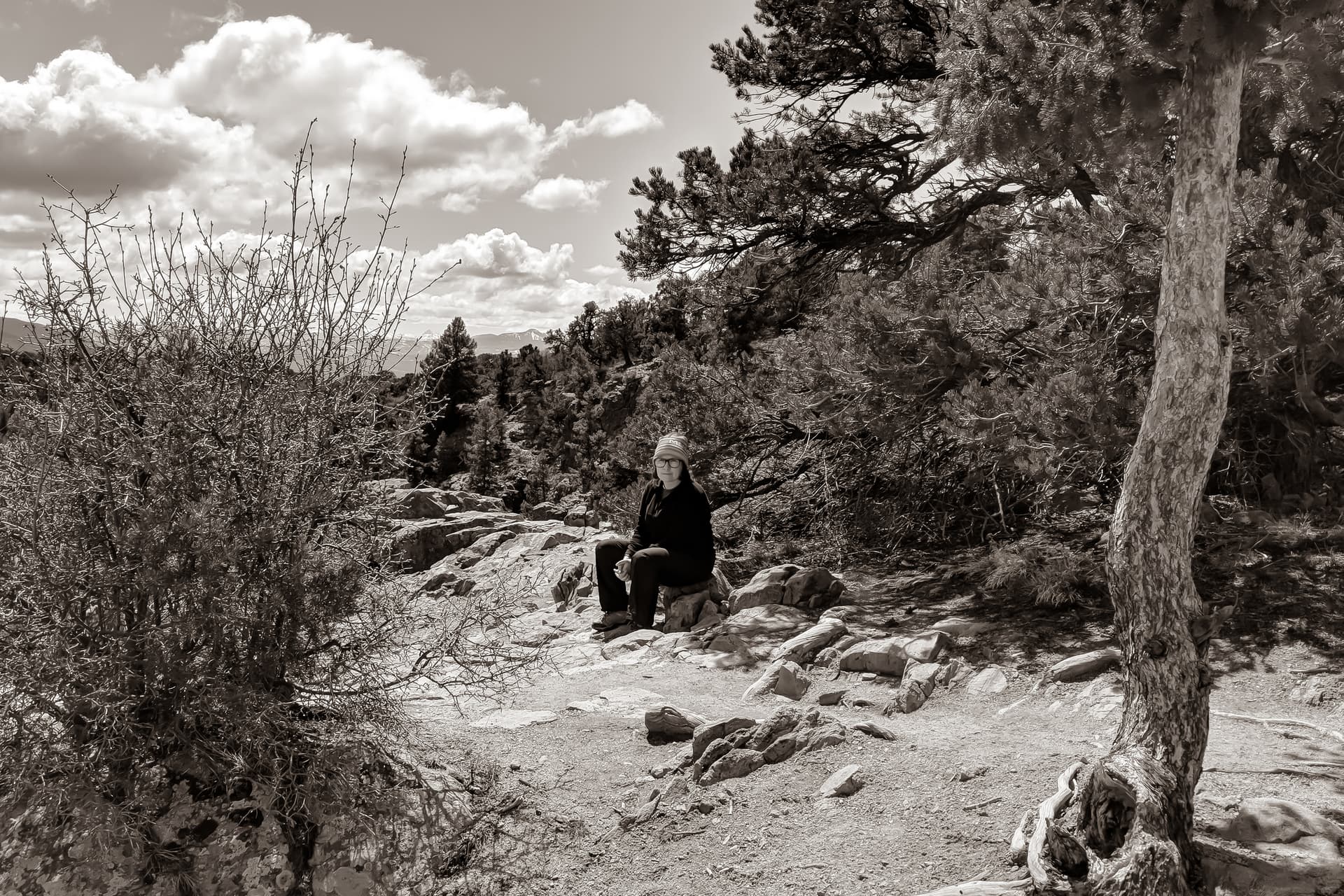 A black-and-white photograph of Len sitting on a rock just outside of a pine and juniper forest.