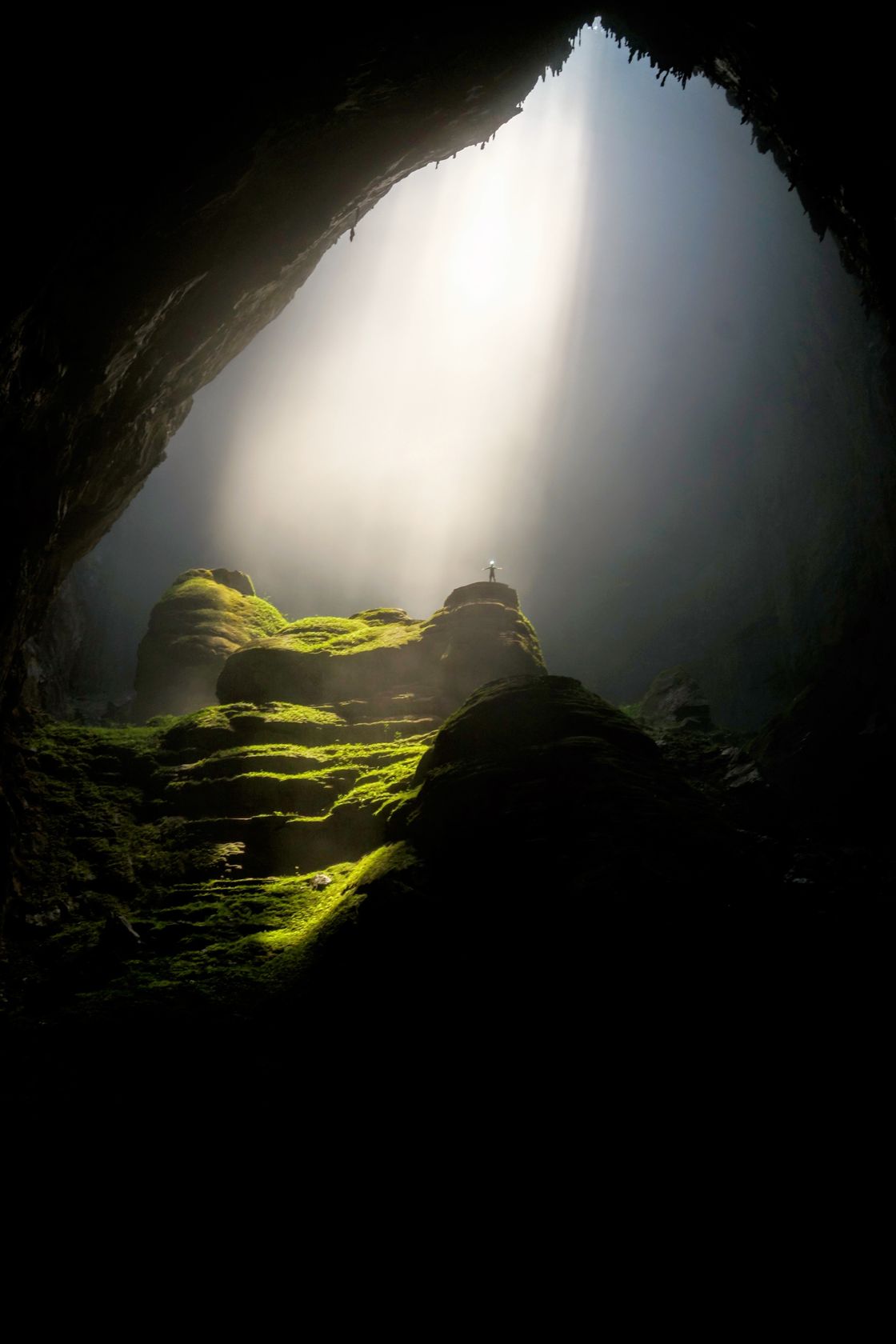 Lonely person standing on moss-covered rocks in a beam of light in a huge cavern.