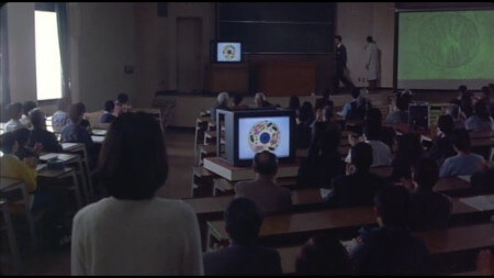 A packed lecture theatre, with Kiyomi looking on, as images of Mitochondria are shown A packed lecture theatre, with Kiyomi looking on, as images of Mitochondria are shown