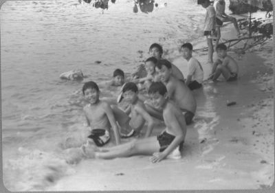 A black and white photo of nine schoolboys sitting on the sand at Punggol Beach close to the waters. Gentle waves lap their legs as they pose for the camera.