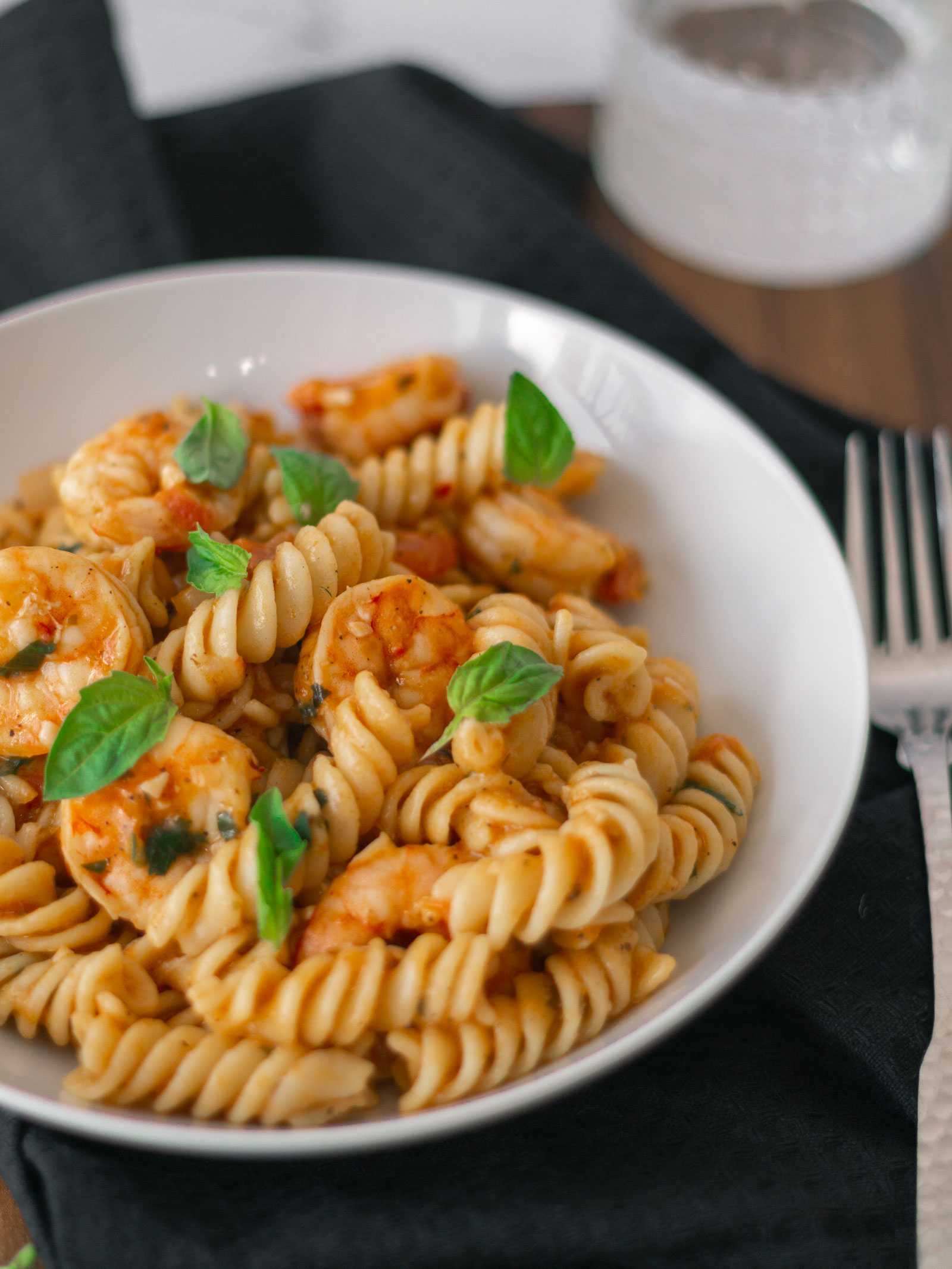 This is an image of Oliver's Tomato Shrimp Pasta. This delicious dish is sitting in a white bowl with fresh oregano sprinkled on top. The al dente pasta, fresh tomatoes, and perfectly cooked shrimp complimented with freshly minced garlic. This may become your summer go-to, as it is light yet filling! 