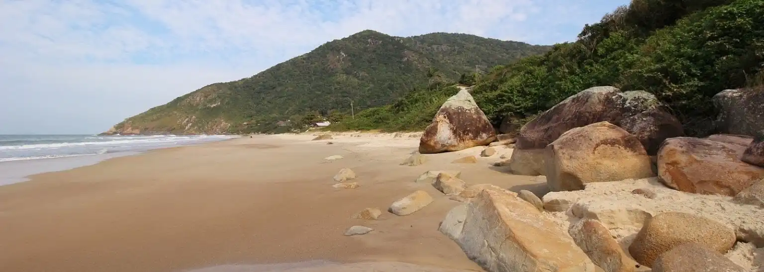 Foto da areia com grandes pedras em tons amarelados e morro ao fundo e pequena faixa de praia a esquerda