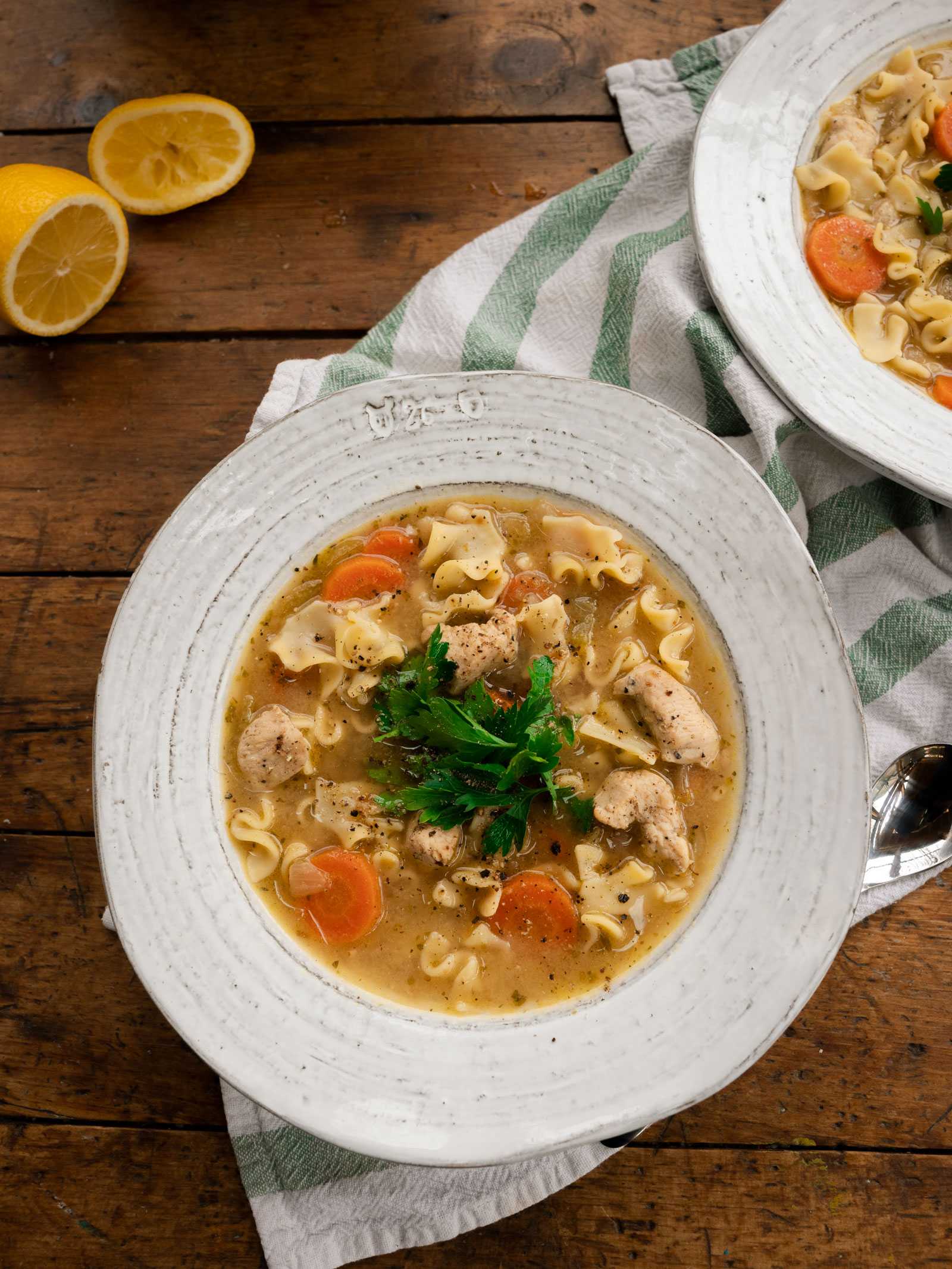Here is a bowl of Oliver-made chicken noodle soup, sitting on a wood harvest table on top of a white & green striped tea towel.  This is a classic recipe that is cozy and delicious. This hearty soup will warm you up from the inside out. It is perfect on its own but also tastes great accompanied with fresh bread.