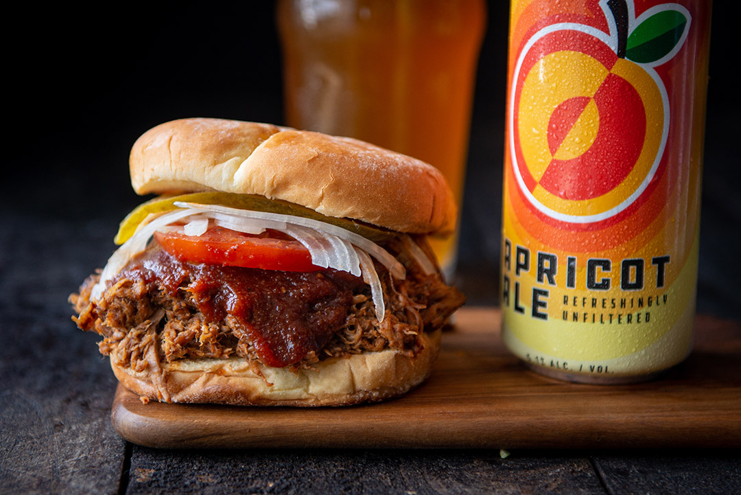 Pulled pork sandwhich on a cutting board next to a can of Apricot Ale