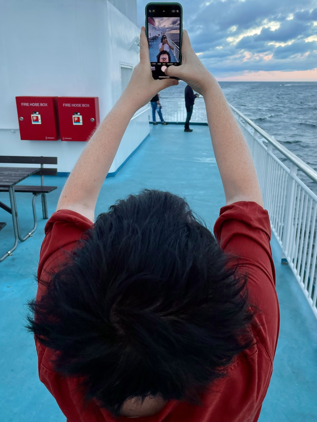 A woman standing on the deck of a ferry, taking a selfie with her phone. The camera app preview shows a man standing right behind her, taking a photo of her taking a photo of both of them.