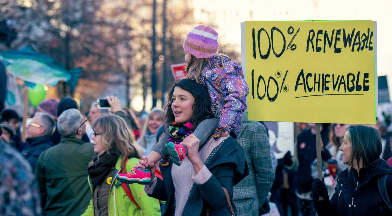 A group of people at a climate change protest. In the centre of the photo is a young woman with a child on her shoulders