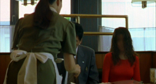 A screenshot of a waitress serving a table with a man in a suit and woman in a red dress without a face. From the film 'Seance'.