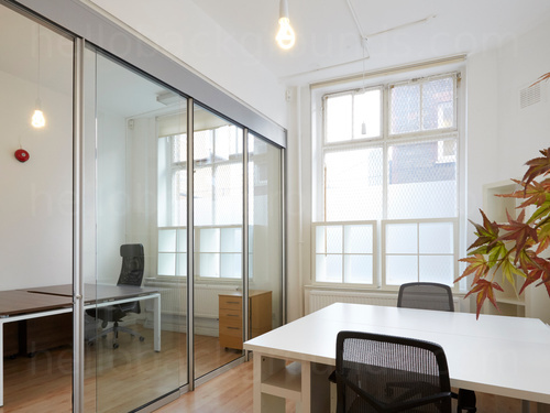 Compact office workspace featuring large white table with two black office chairs and glass walls with view into next office Google Meet background
