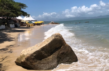 Grande pedra na area com onda batendo levemente e faixa de area atrás mostrando um bar ao fundo com alguns guarda-sóis