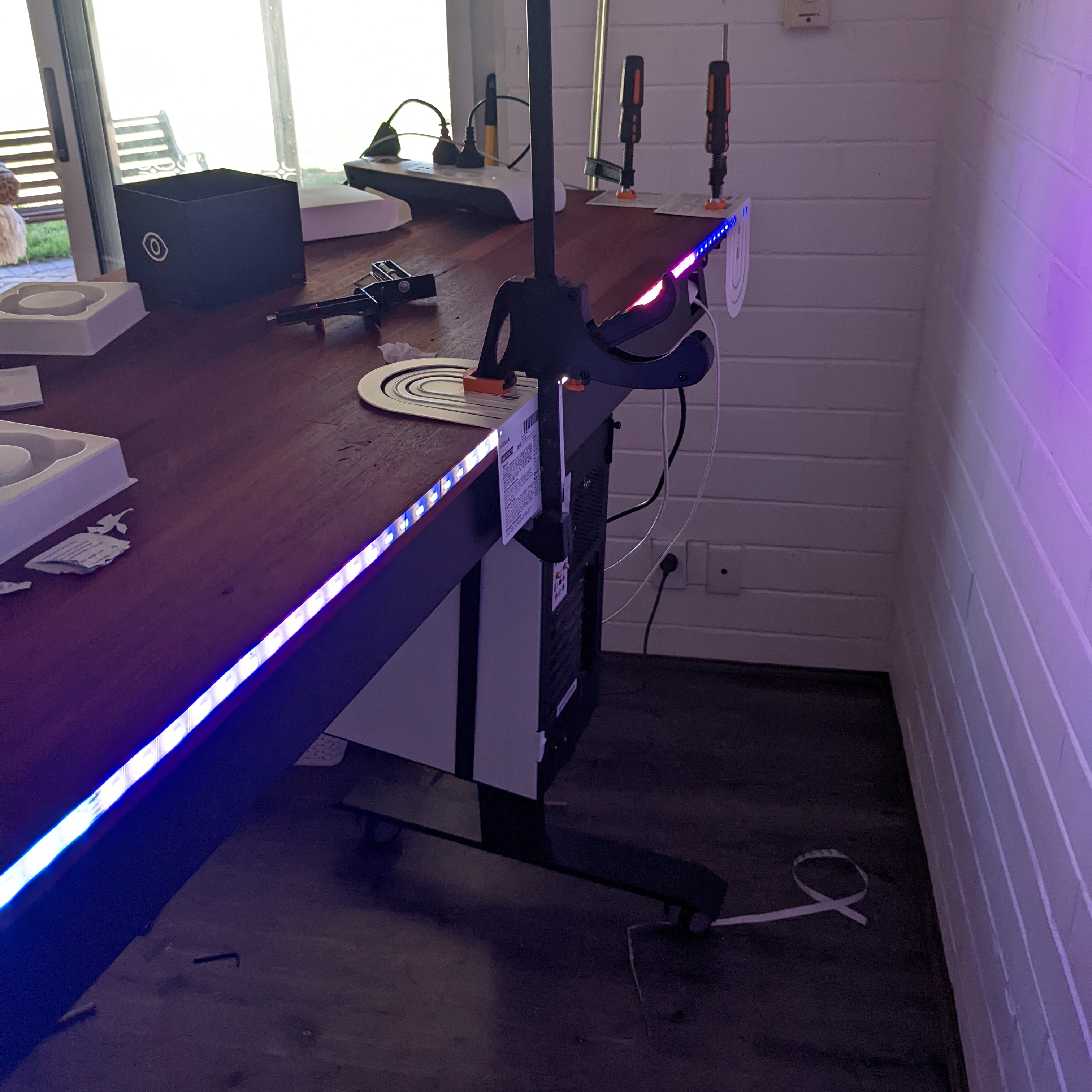 LED light strip attached to the back of the desk top, with a bookend being used to hold it to the back of the desk and a clamp being used to hold the bookend in place, in the background there are several other clamps doing the same thing