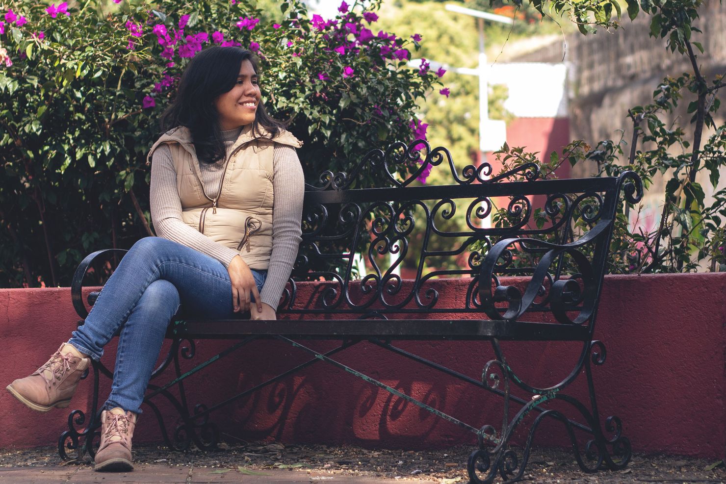 estrella, wearing jeans and a puffy vest, sits smiling on a black metal bench in front of a flowering bush