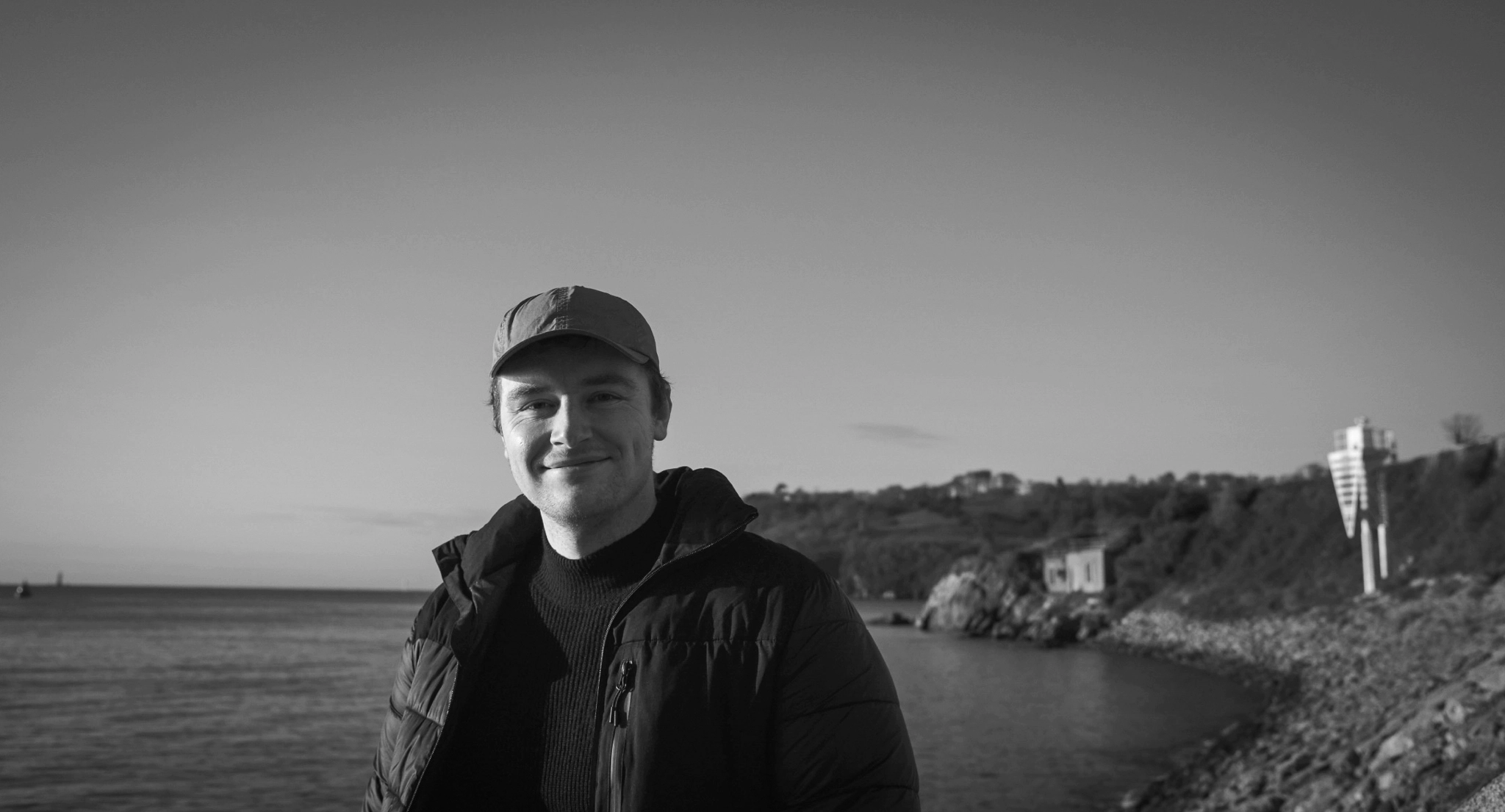 A photograph of me smiling at the camera, taken on a cold, sunny day on an outlook over the ocean. Plymouth, February 2022.