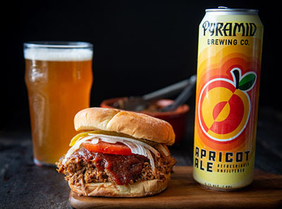 Pulled pork sandwhich on a cutting board next to a can of Apricot Ale