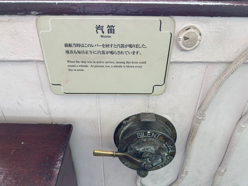 A photograph of a brass lever with a few rotary setting. The detailing on the housing shows one of the labels to be “silent”. A sign above the lever reads: “When the ship was in active service, turning this lever could sound a whistle. At present, too, a whistle is blown every day at noon.”