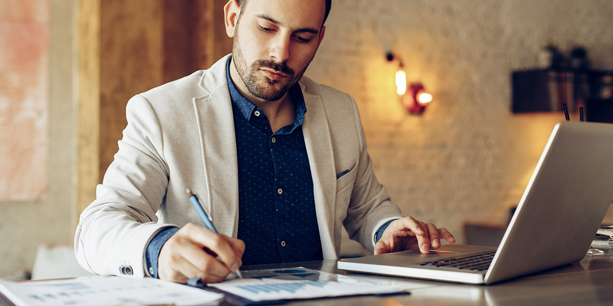 What is the CONCATENATE function in Excel? How can you use it? A man sitting at a desk, writing something down, with a laptop in front of him