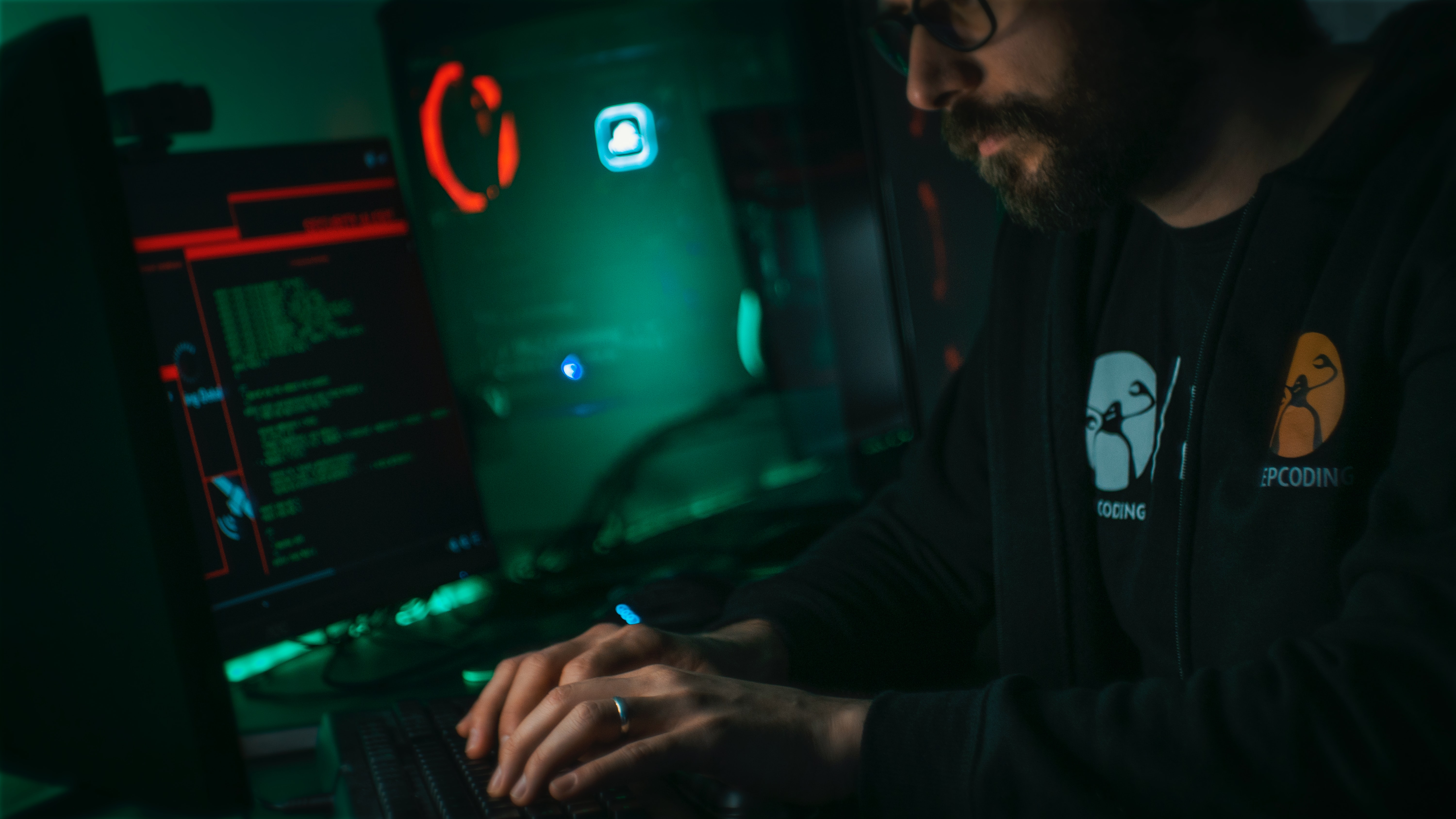 A man sits in a dark room facing his computer, working on cybersecurity coding.