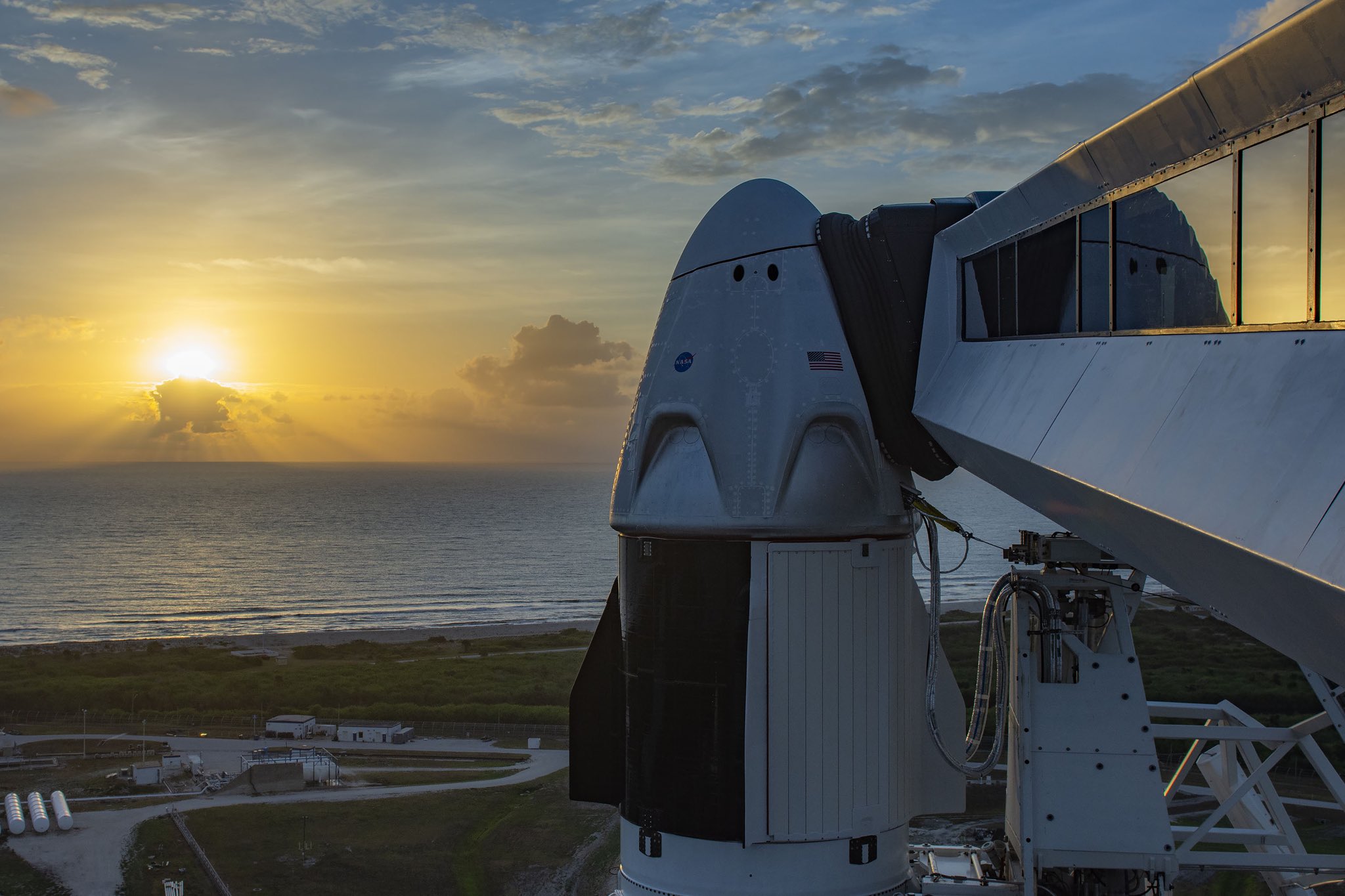Imaginea 1: Capsula Dragon2 (sau Crew Dragon), așteptând o zi cu un cer mai senin, pe rampa de lansare LC-39A de la Kennedy Space Center. În drapta imaginii se poate observa brațul folosit pentru accesul echipajului la capsulă. (Sursa foto: SpaceX)