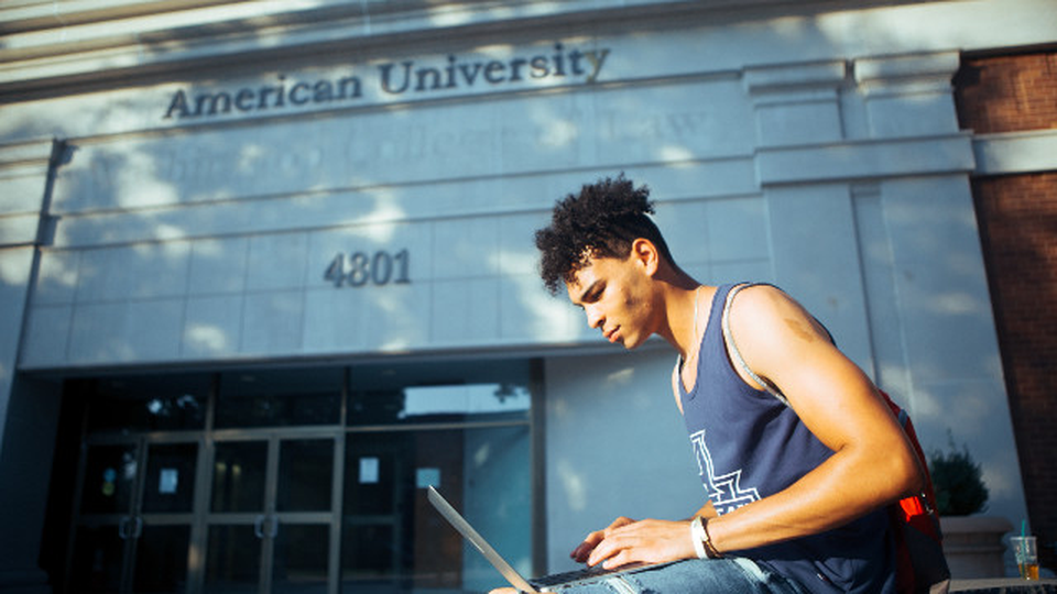 A man seated in front of an American University building typing something into a laptop