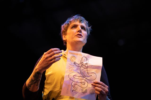 Miriam holding a paper with a drawing of
a half-child, half-serpent,
gesturing and talking
