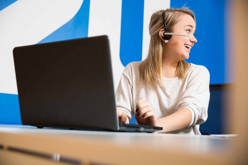 Woman laughing on a wireless headset and typing on a laptop in front of a blue wall with the 2U logo on it