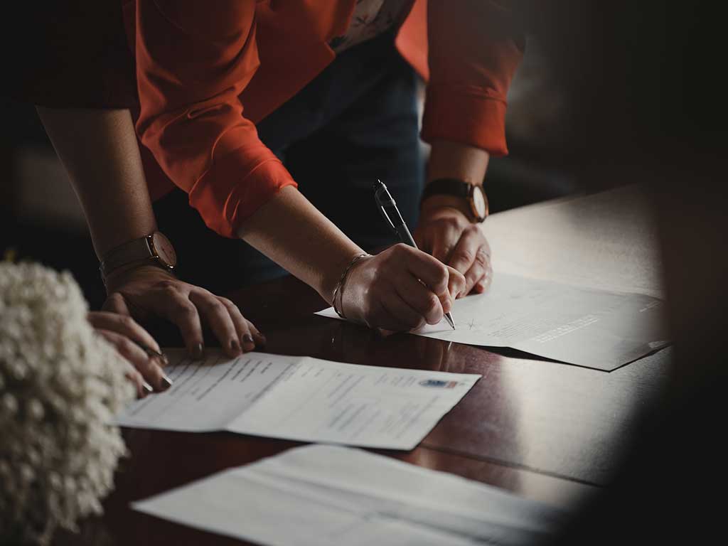 Someone signing a document on a table with a pen while someone else witnesses, Photo by Romain Dancre on Unsplash