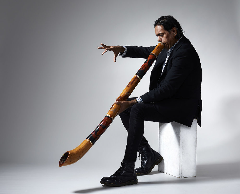 William Barton playing the didgeridoo on a stool in studio with a white backdrop and harsh studio flash lighting