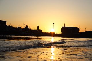 Sun rising behind Shoreham power station and Shoreham lifeboat station. A small fishing boat causes the water to ripple in the foreground.