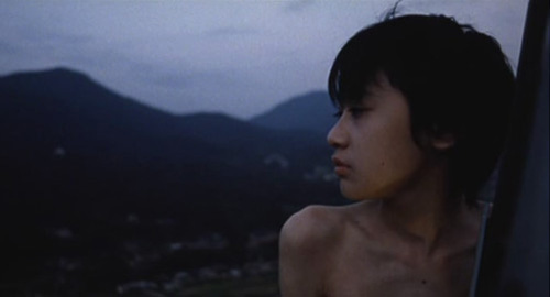 A close-up screenshot of young boy standing from tall height looking off into the distant mountains. From the film 'Antenna'.