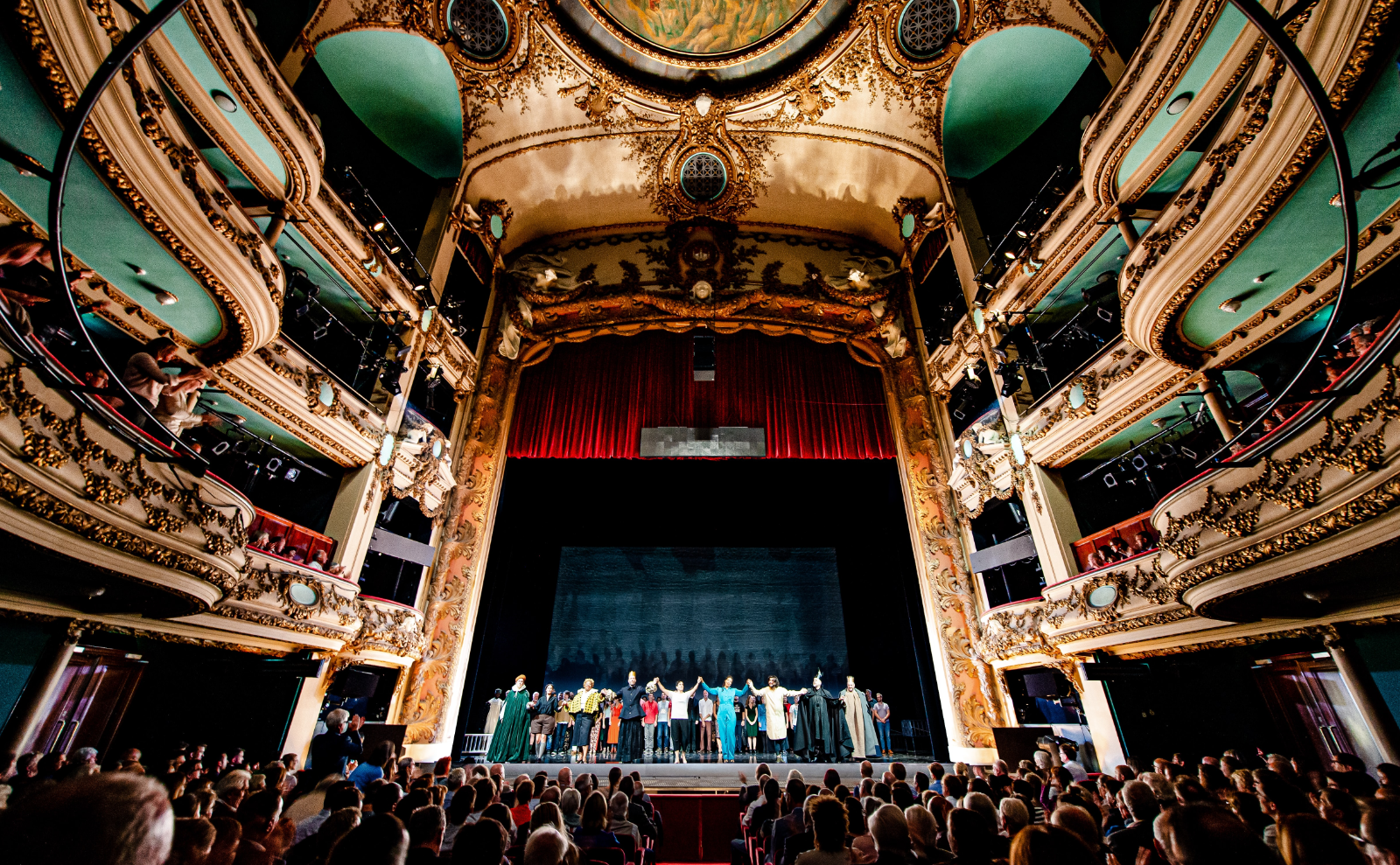 baroque theater with metallic gold balconies and a full audience sitting in red velvet chairs, on the stage is a cast holding hands to take a curtain call