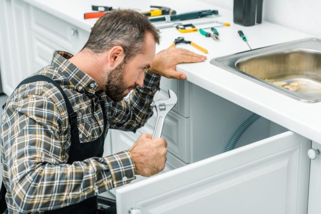 repairman holding adjustable wrench and looking under broken sink in kitchen