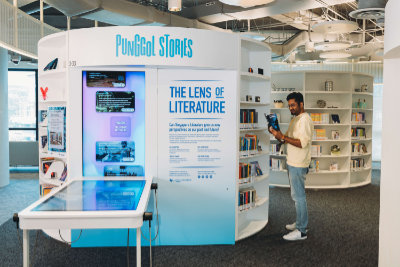 A photo of the Lens of Literature kiosk, that has a multimedia table fixed to it. Next to the kiosk is a bookshelf, with a man reading a book.