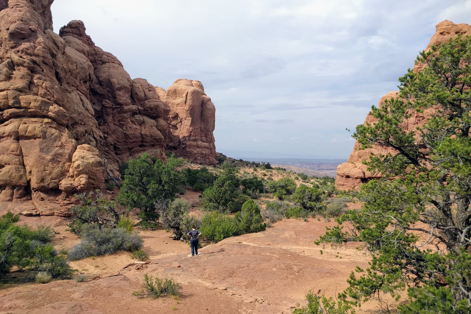 Len, in a gray shirt, stands far down a trail of red sandstone, her hands on her hips. Walls of red rock stand to her left and right, and a small grove of juniper trees is in front of her.