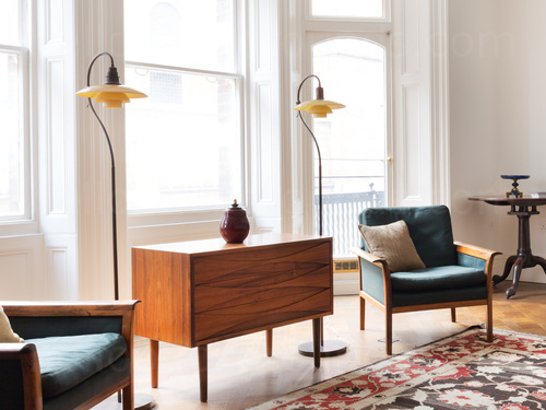 Large airy living room featuring two wooden chairs in dark green upholstery and wooden cabinet in front of 3 large windows  Zoom background