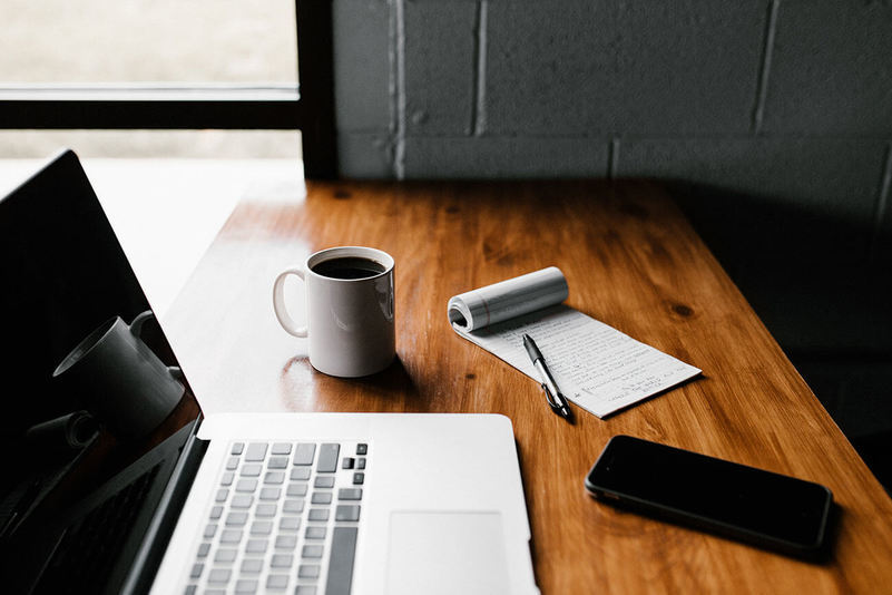 Uma mesa com alguns utensílios em cima: um notebook, uma xícara de café, um celular e um bloco de notas com uma caneta em cima