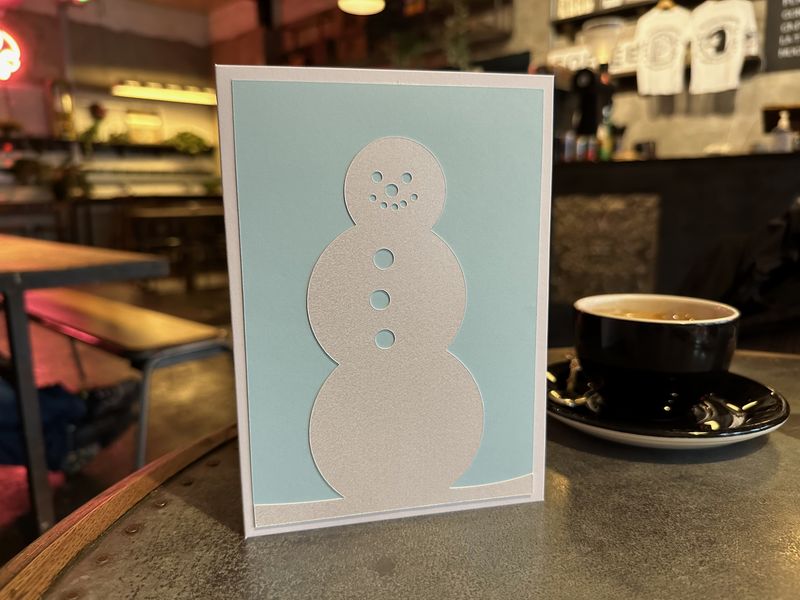 A hand made Christmas card standing on a table in a cafe, next to a cup of coffee. The card is white, with a blue panel on the front. On top of the panel is a snow man cut from white card with a specular snow shine to it.