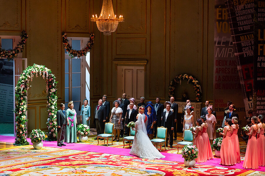 Une scène d'opéra. Au premier plan, un tapis de style Empire, rouge avec des motifs jaunes et bleus. Dessus, un couple de mariés s'avance en direction d'une arche fleurie.