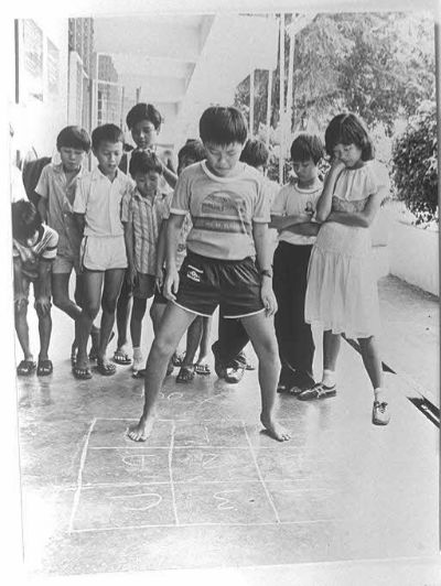 A boy plays hopscotch in a corridor while several other children look on from behind him. The hopscotch tiles have been drawn on the floor in chalk.